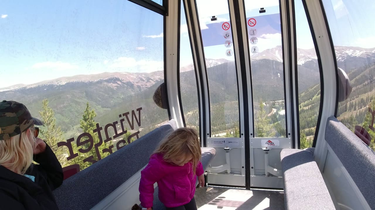 The young girl stands near the gondola door in Winter Park, Colorado while the seated woman looks toward her and mountain slopes fill the background.
