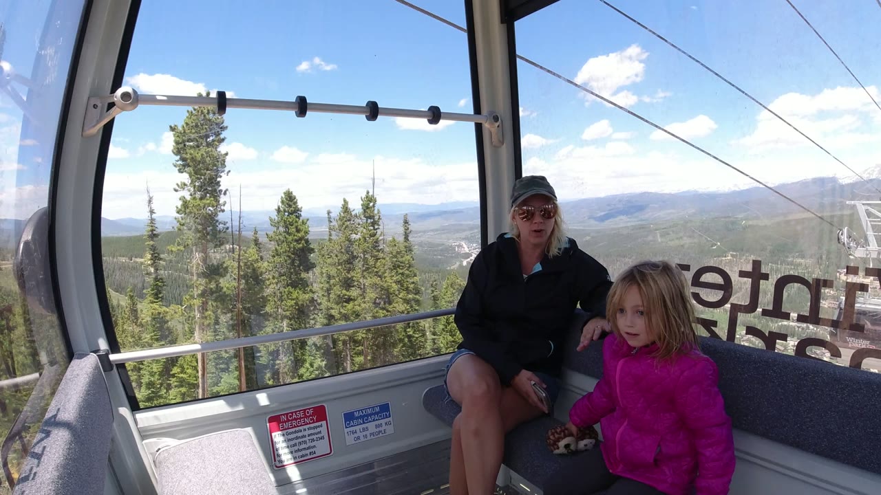 A woman in sunglasses and a young girl in a pink jacket sit inside a Winter Park, Colorado ropeway cabin, with pine trees, cable lines, and distant hills visible through the windows.