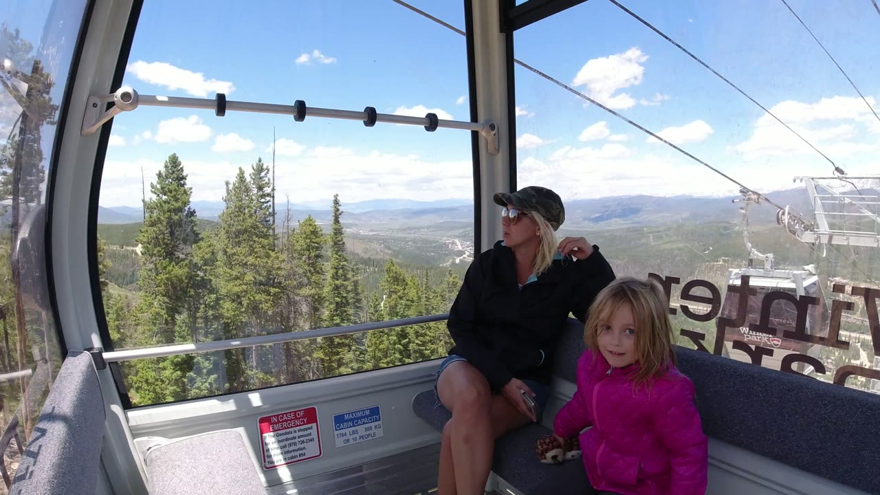 Inside the Winter Park ropeway cabin, the woman turns toward the window while the child in a bright pink jacket faces forward, with forested hills and lift cables beyond the glass.