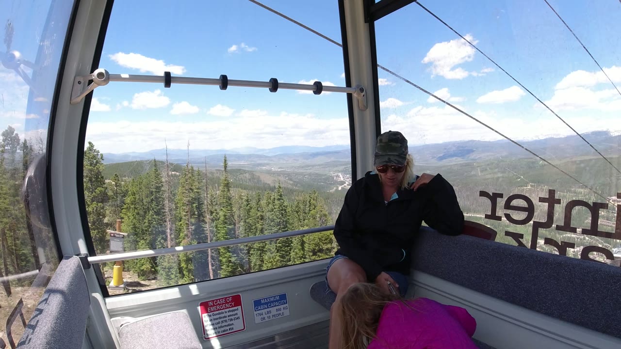 Inside a Winter Park, Colorado cable car, a seated woman in sunglasses and a cap looks down while a child in a pink jacket leans near the window and forested slopes fill the background.