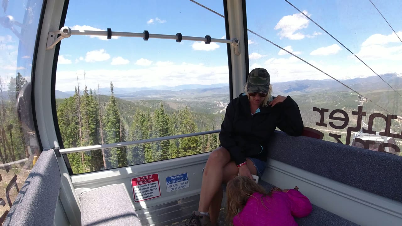 A second moment inside the Winter Park gondola shows the woman seated near the right window, the child lower in frame, and a wide view of evergreens and distant hills through the glass.