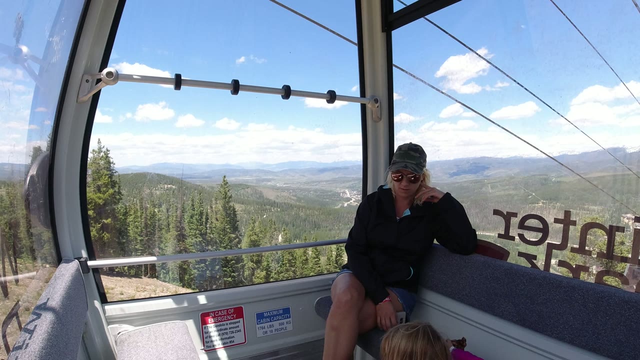 A woman sits inside a Winter Park gondola cabin while a young child is partly visible at the bottom edge and evergreen mountains fill the window view.