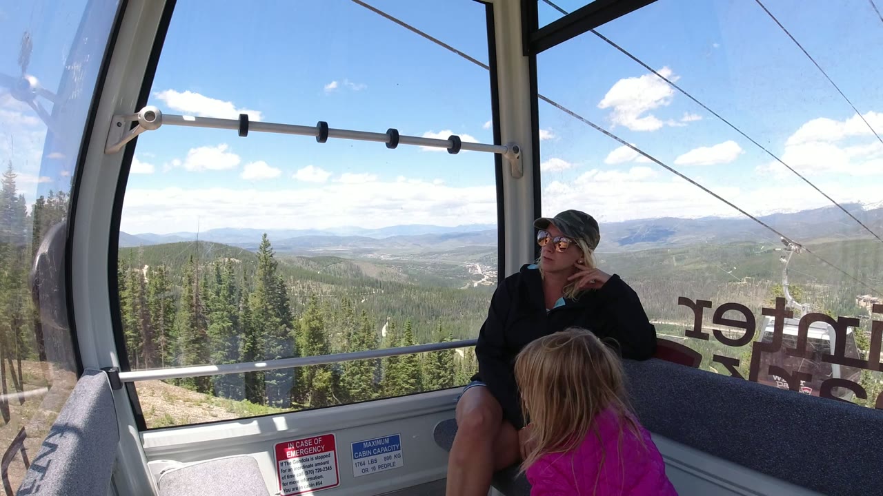 The woman remains seated in the Winter Park ropeway cabin as the child in a bright pink jacket appears lower in frame against the wide mountain window.