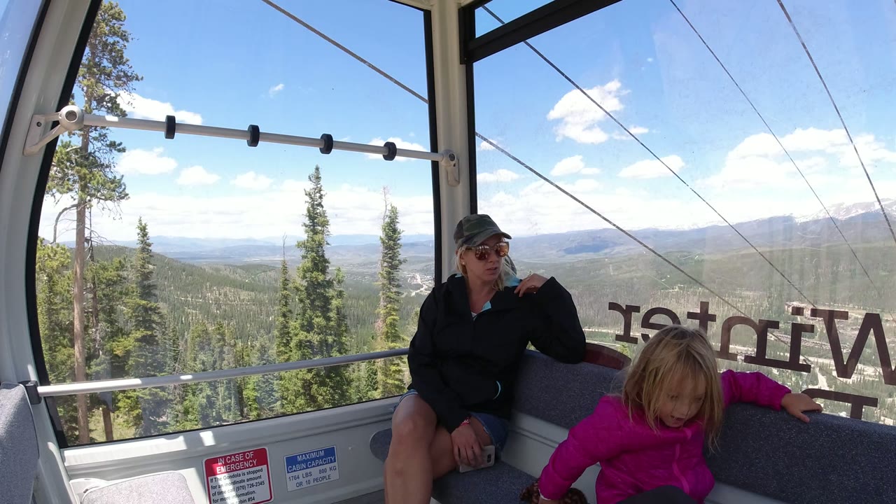 Inside a Winter Park, Colorado gondola, a woman in sunglasses and a child in a pink jacket sit by large windows overlooking evergreen trees and distant mountains.