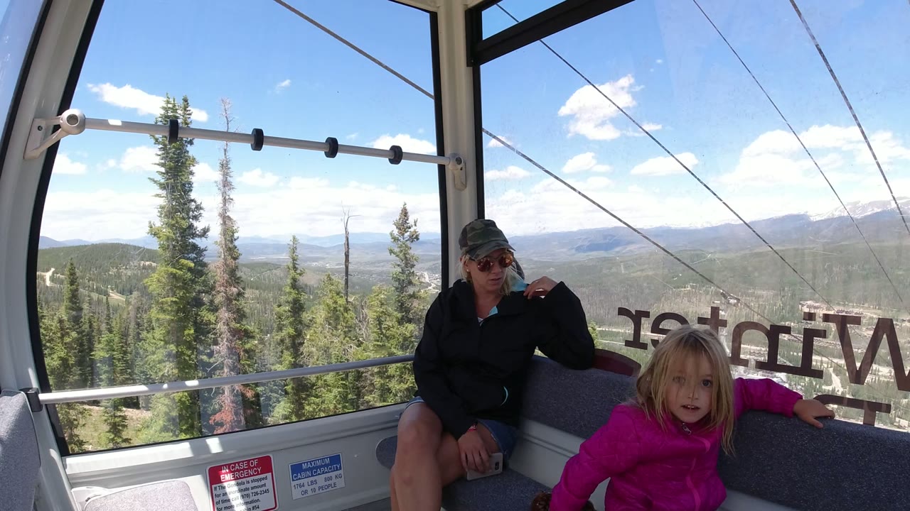 A child in a bright pink jacket looks toward the camera inside the Winter Park gondola as a woman sits behind her and cable lines cross the mountain view outside.
