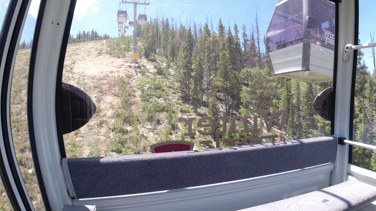 Winter Park gondola interior with a passing cabin near the upper right, a lift tower ahead, evergreen trees on the hillside, and no visible people.