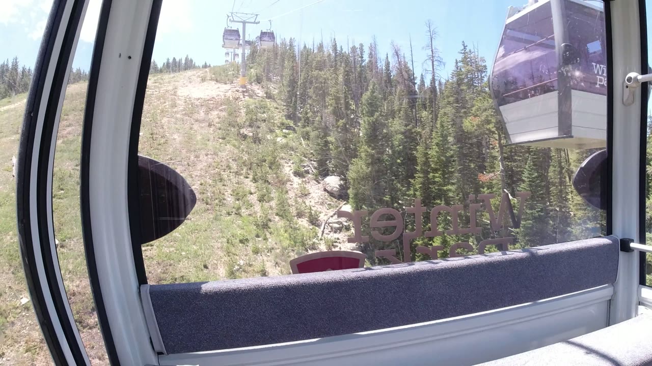 A darker gondola car passes near the right side of the Winter Park window view, with evergreen trees and pale rocky ground behind it, and no visible people.