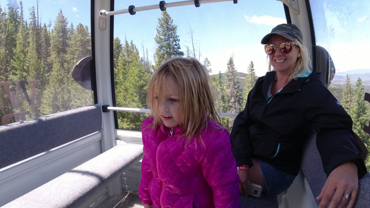 At 0:00, a child in a bright pink jacket sits near the window of a Winter Park lift cabin while a woman in sunglasses sits behind her and evergreen trees fill the view outside.