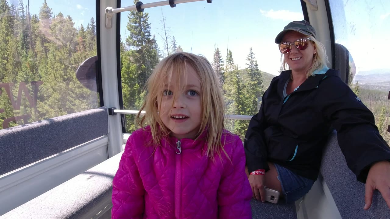 At 0:01, the child faces the camera more directly inside the Winter Park lift cabin, with the woman seated to the right and sunlit forest visible through the windows.