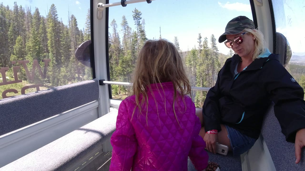 Child in pink jacket standing beside a seated woman inside a mountain lift cabin.