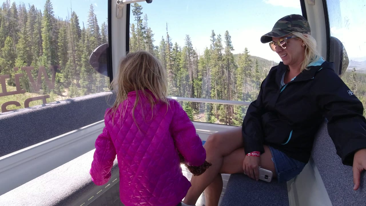 A girl in a pink jacket faces the window of a Winter Park, Colorado lift cabin while a woman in a dark jacket sits on the opposite bench, with trees and hills visible outside.
