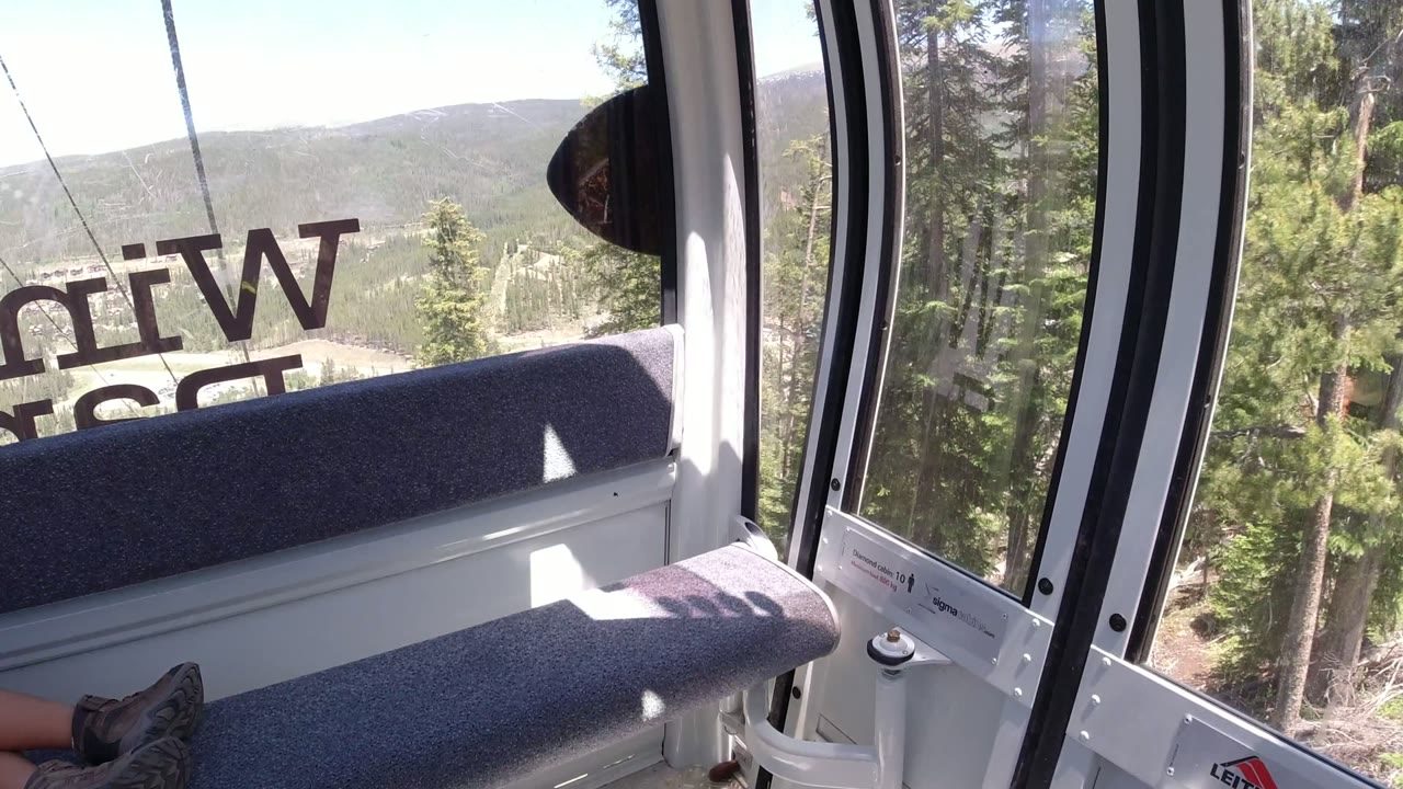 A Winter Park, Colorado cable car interior looks out over dense green pines and hazy hills, with the bench backlit by bright sun and no visible faces.