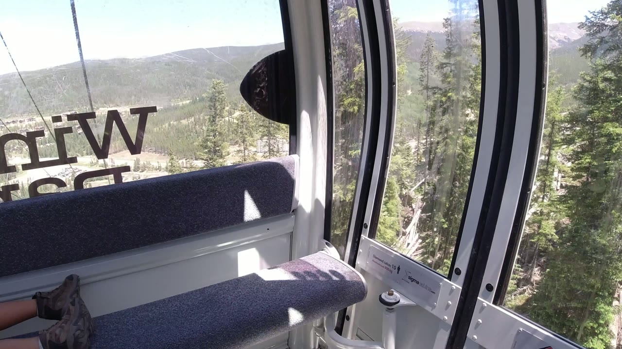 A Winter Park, Colorado cable car window view shows the gray bench in the foreground, reversed lettering on the glass, evergreen forest below, and hazy mountain ridges beyond.