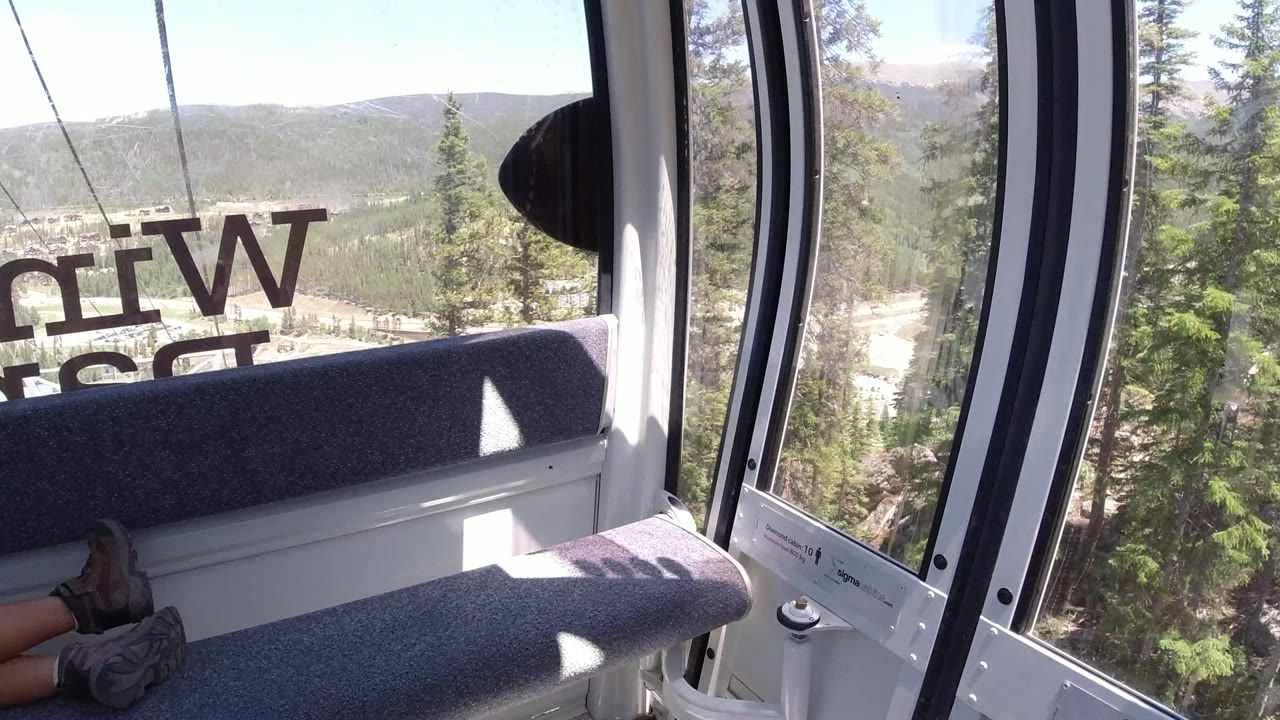 Inside a Winter Park, Colorado cable car, gray bench seats and reversed window lettering frame evergreen trees and a distant mountain valley, with partial passenger legs visible at the lower left.