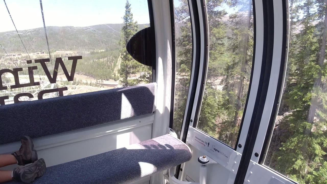 A Winter Park cable-car cabin interior shows gray seats, dark window trim, reversed glass lettering, nearby evergreens, and mountain ridges beyond, with passenger shoes partly visible.