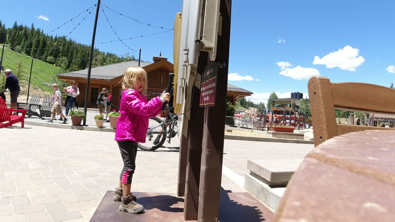 Child in a pink jacket standing beside a post in an outdoor mountain plaza.