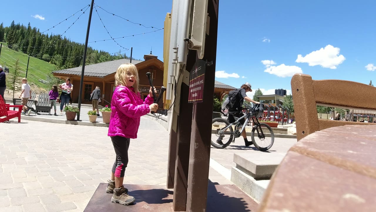 The child in the bright pink jacket turns more toward the camera in Winter Park, Colorado, while a cyclist passes behind her and red chairs and pine-covered slopes remain visible.