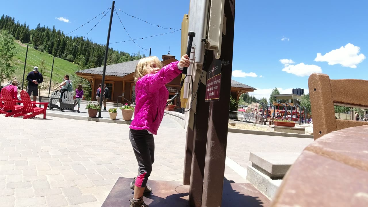A young girl in a bright pink jacket stands beside a doorway in Winter Park, Colorado, with red chairs, visitors, green slopes, and pine trees visible behind her.