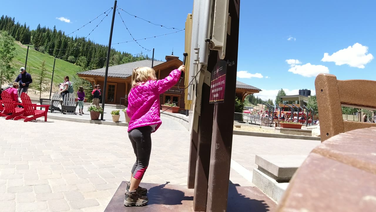 The girl turns partly away from camera and extends the stick toward the wall or doorframe in Winter Park, Colorado, while visitors and red seating remain in the plaza behind her.