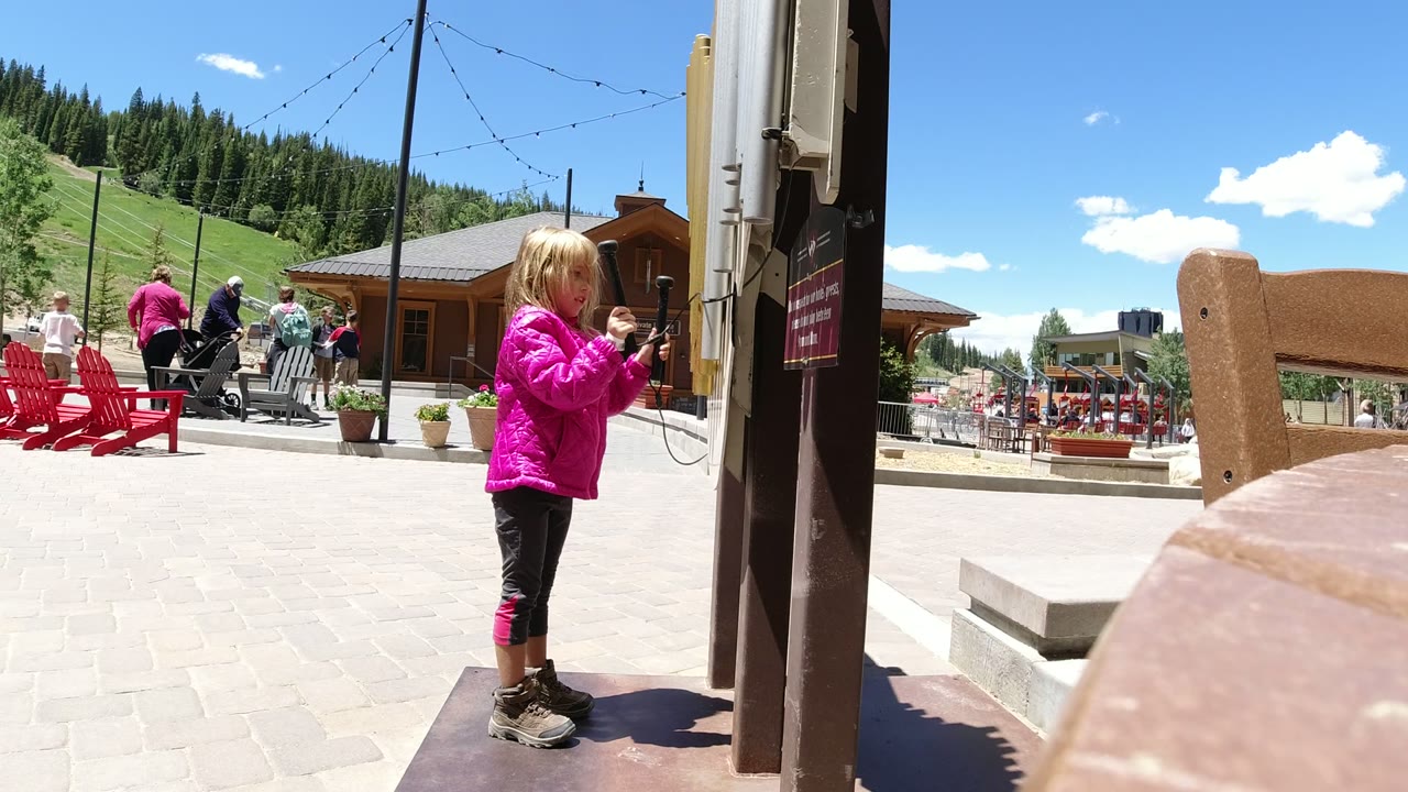 A young girl in a bright pink jacket stands near a dark post and hanging cord in a Winter Park, Colorado plaza, with red chairs, adults, wood buildings, green slopes, and pine trees behind her.