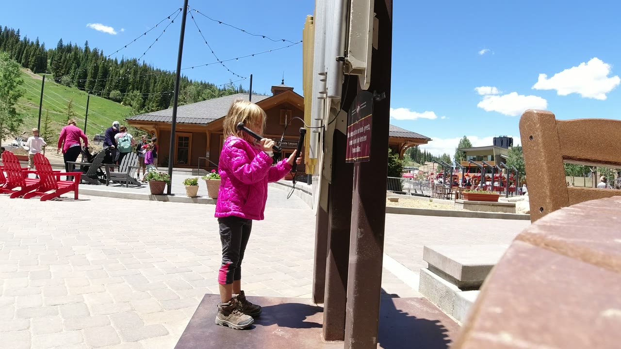 The girl looks down while holding the cord near the post in Winter Park, Colorado, with empty red chairs, background visitors, a wood building, and green mountain slope visible.