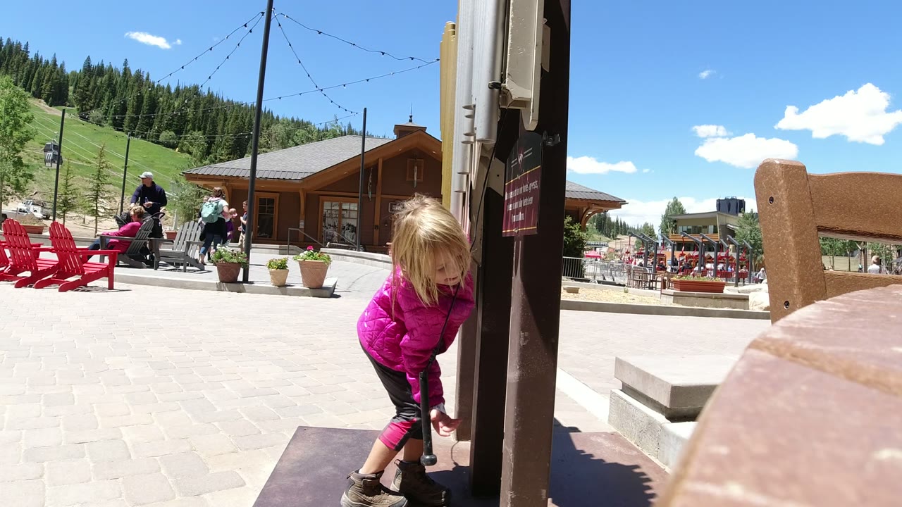 Young blonde girl in a pink coat bends beside a public instrument post in a sunny plaza.