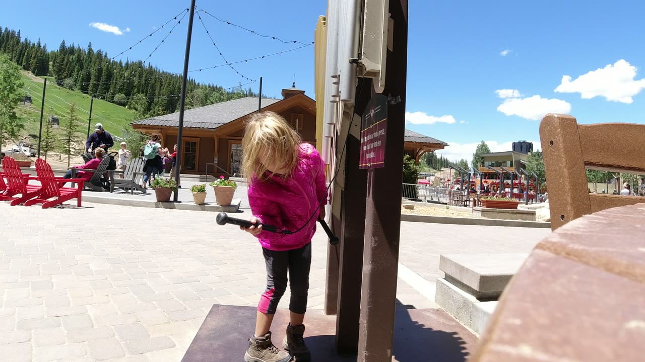 At 0:01 in Winter Park, Colorado, the girl stands more upright beside the vertical post, with her face partly obscured by hair and the red chairs, plaza visitors, and treed slope in the background.