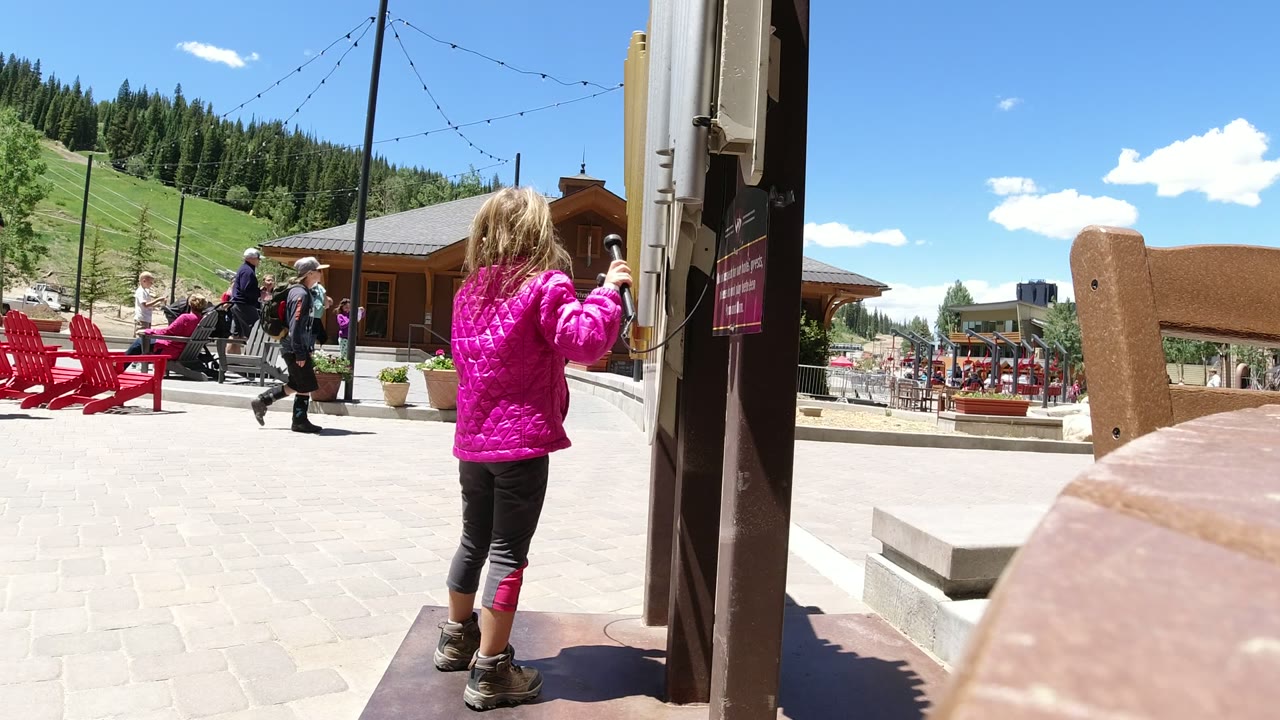 Girl in a pink jacket stands by a sign post in a sunny plaza as people walk behind her.