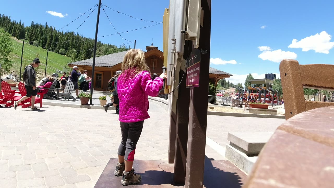 At 0:01 in Winter Park, Colorado, the girl remains beside the sign post as a man and seated visitors appear near red chairs in the background.