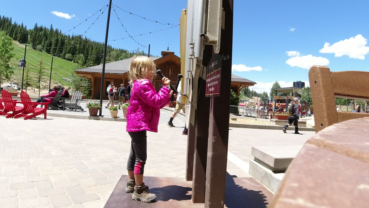 A child in a bright pink jacket stands near a public phone post in Winter Park, Colorado, with red chairs, pedestrians, green slopes, and lodge-style buildings behind her.