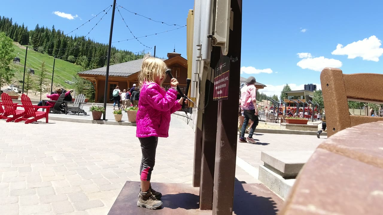 The child faces the phone post in Winter Park, Colorado, with her hands near the handset area as visitors and red chairs remain visible across the paved plaza.