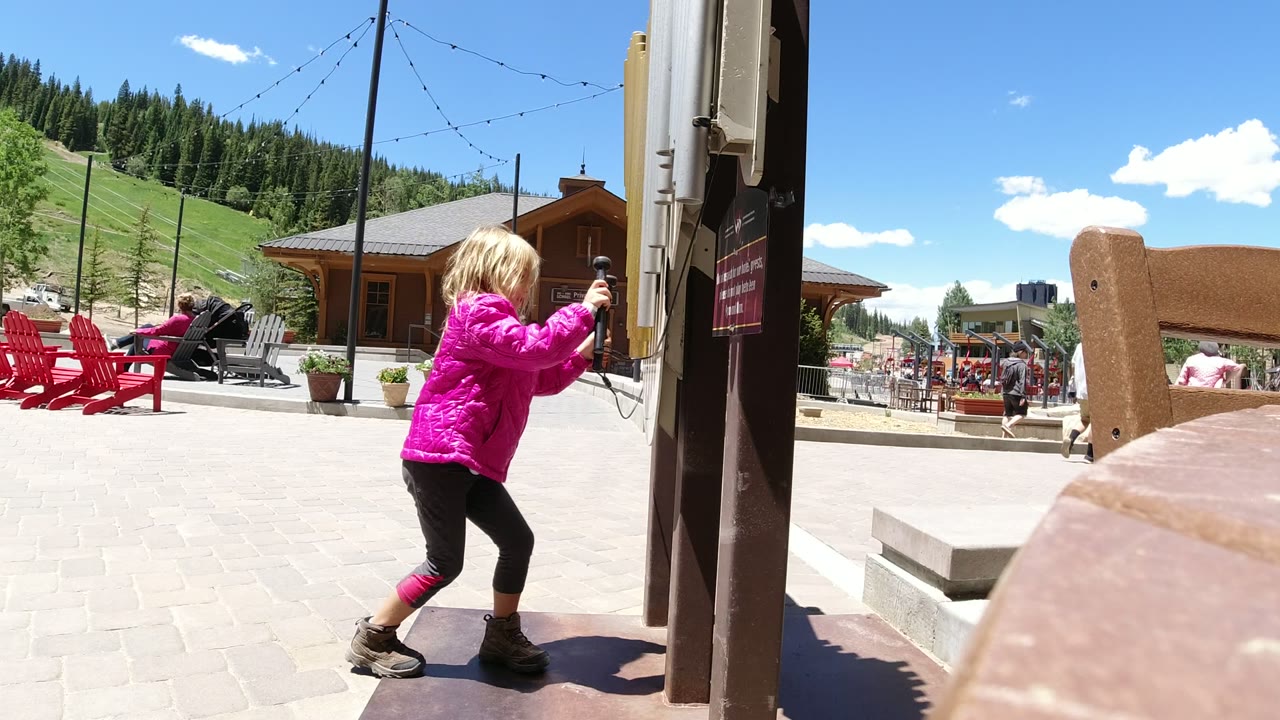 A young blond girl in a pink jacket holds a cord beside a dark post in Winter Park, Colorado, with red chairs, lodge buildings, green hillside, and pine trees behind her.