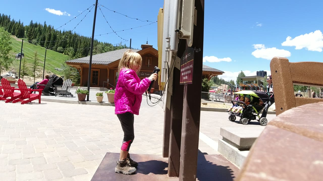 Blonde girl in a pink jacket holding a corded handset beside a post in an outdoor plaza.
