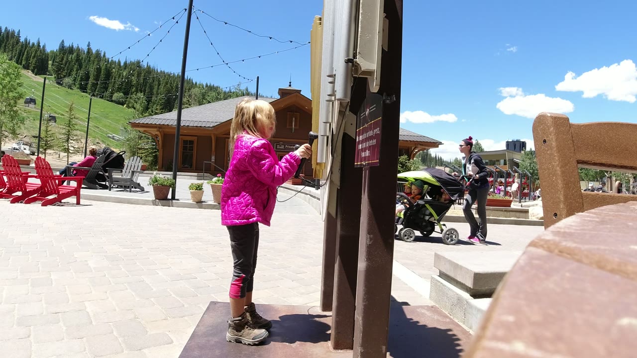 The girl stands side-on at the post in Winter Park, Colorado, holding the handset while an adult with a stroller moves through the sunny plaza behind her.