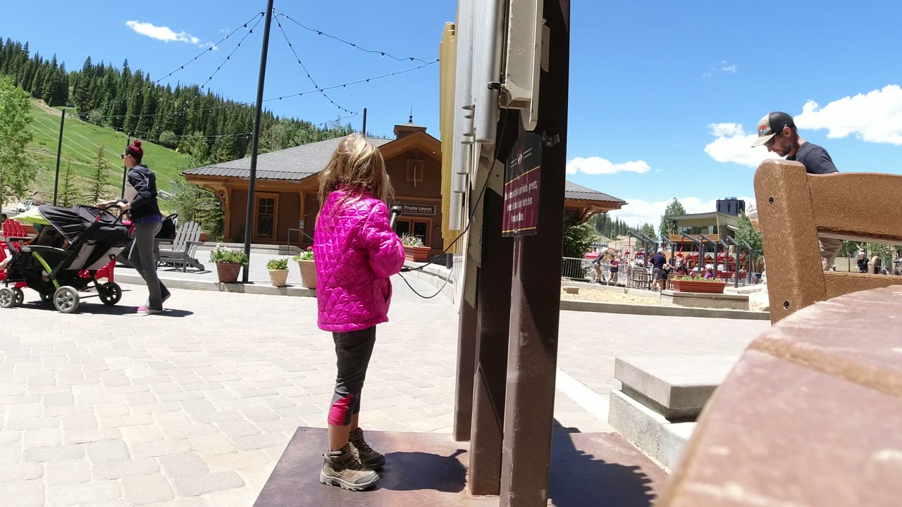 Young girl in a pink jacket stands beside a post in an outdoor plaza.