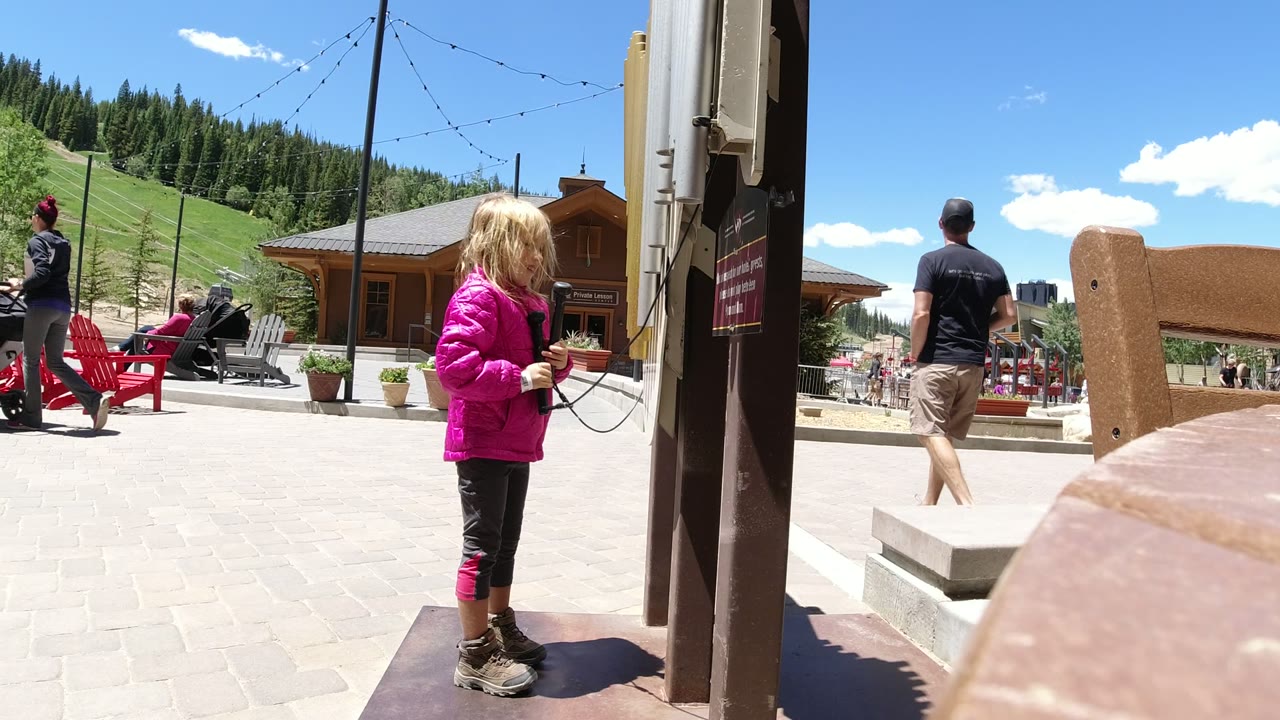 The girl in the pink jacket faces the post while holding the phone cord, with outdoor seating, people, a wooden building, and a forested hill in Winter Park behind her.