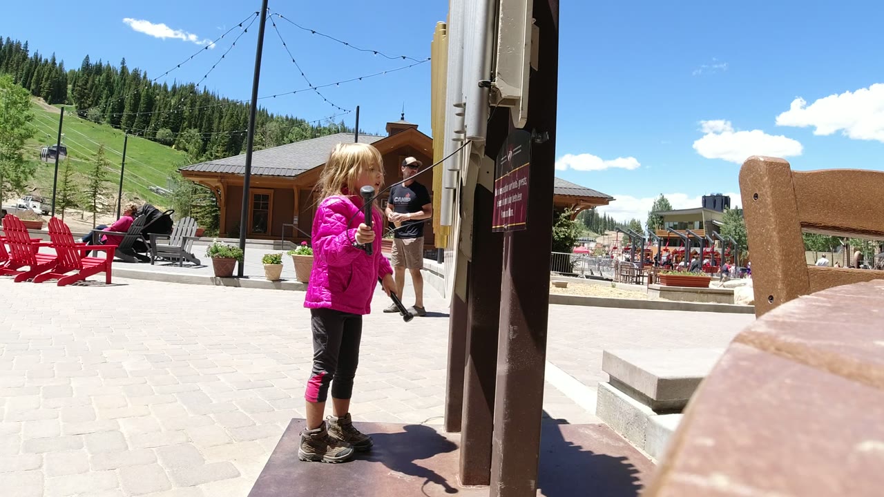 Girl in a pink jacket holding a rope beside a wooden post in an outdoor plaza.