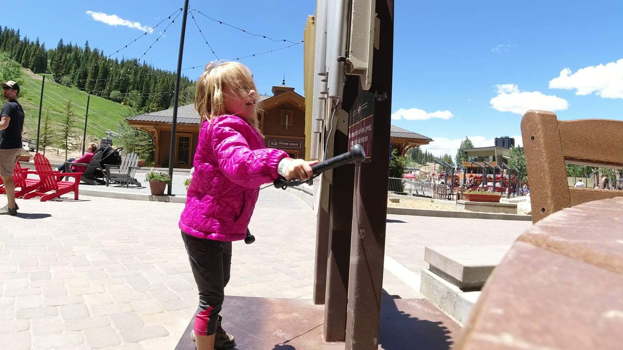 The child turns slightly while holding a black tethered handle beside the activity board, with a paved patio, red chairs, and mountain slope visible in Winter Park, Colorado.