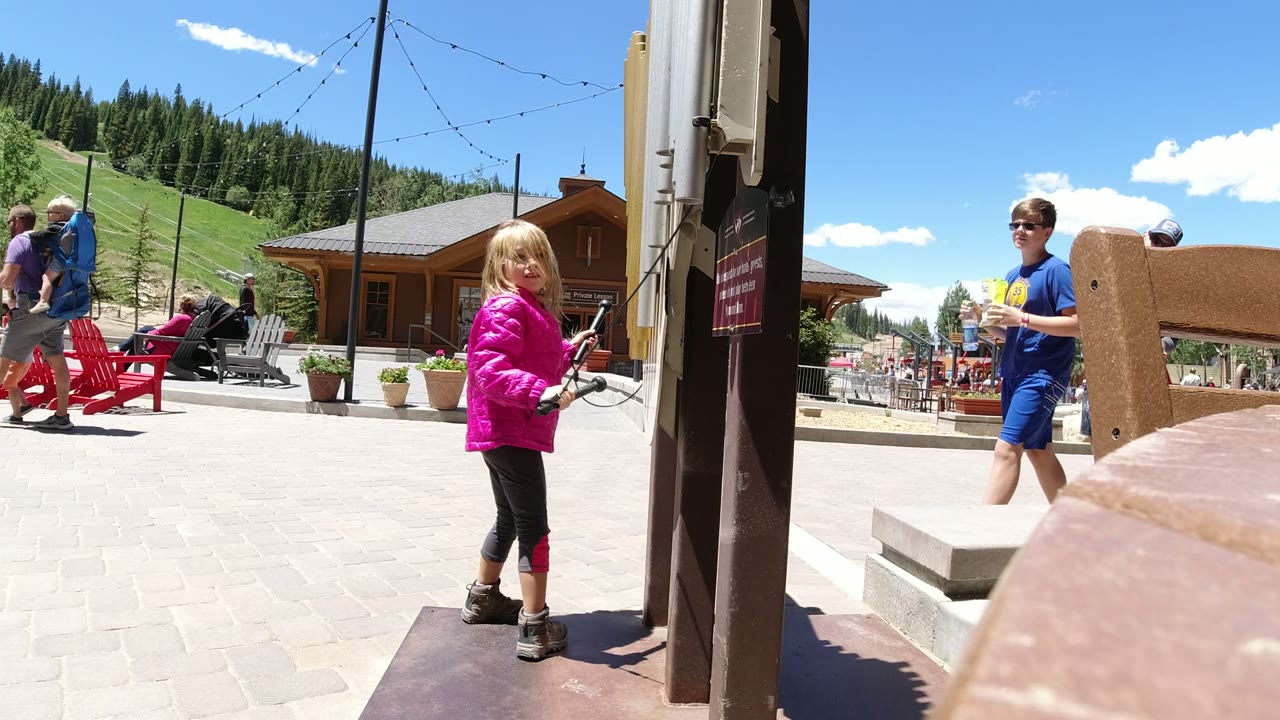A young girl in a pink jacket stands near a wooden post in Winter Park, Colorado, with red chairs, a lodge-style building, green slopes, evergreen trees, and several people in the background.