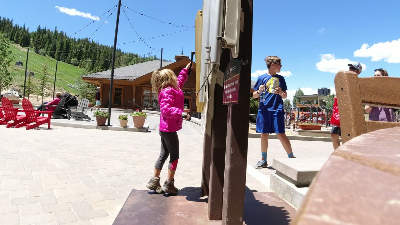 The girl in the pink jacket reaches up near the wooden post in Winter Park, Colorado, while a boy in blue and other visitors move through the sunlit plaza behind her.