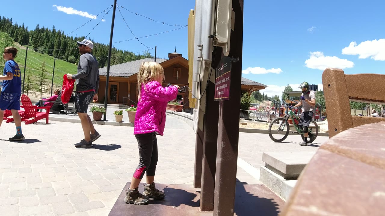 The child moves closer to the xylophone in Winter Park, Colorado, with red patio chairs, a lodge-style building, and a grassy slope behind her.