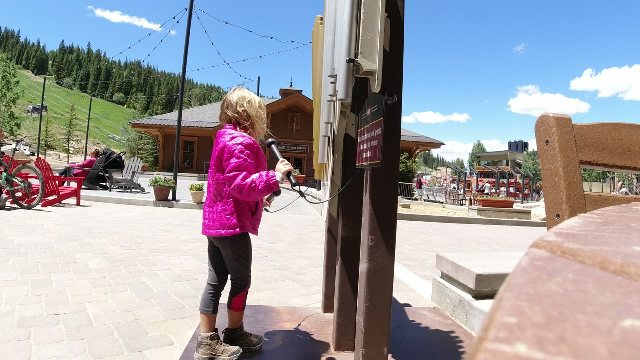 Young girl in a pink jacket standing beside a dark metal structure outdoors.