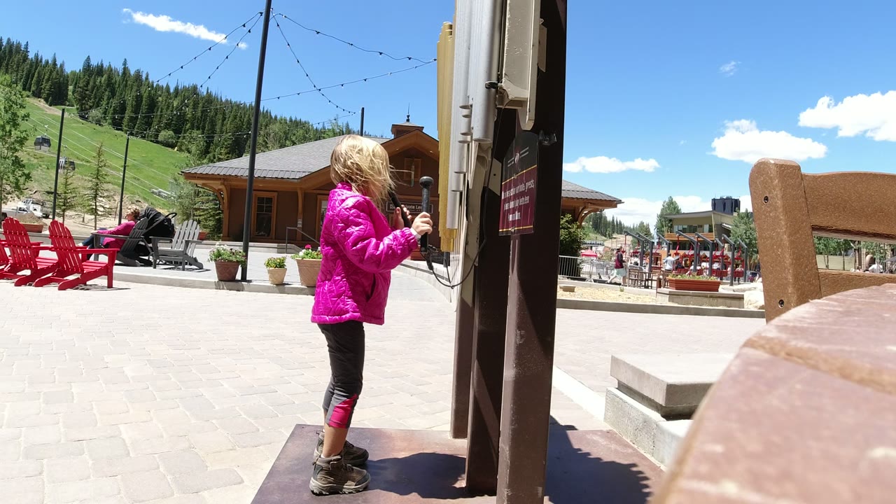 The girl faces the metal wall or post more directly in Winter Park, Colorado, holding a small object or cord while the plaza seating and hillside remain visible behind her.