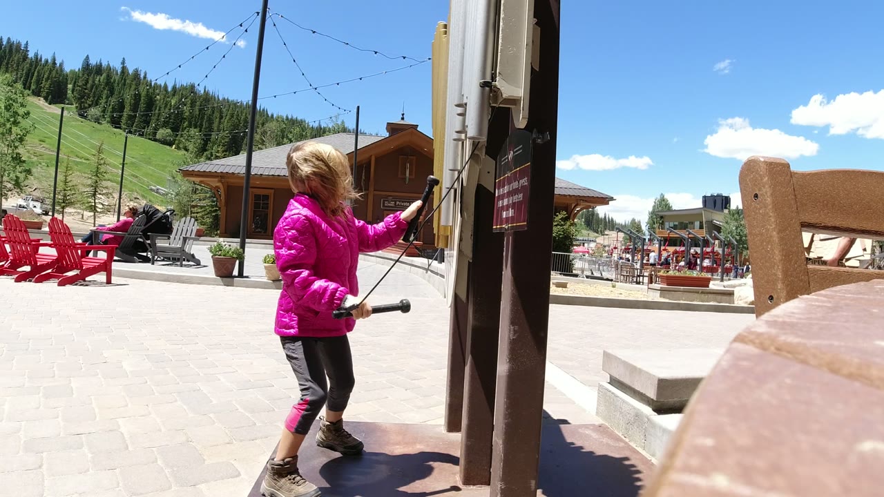Young girl in a pink jacket tapping a sideboard with sticks near an outdoor plaza.