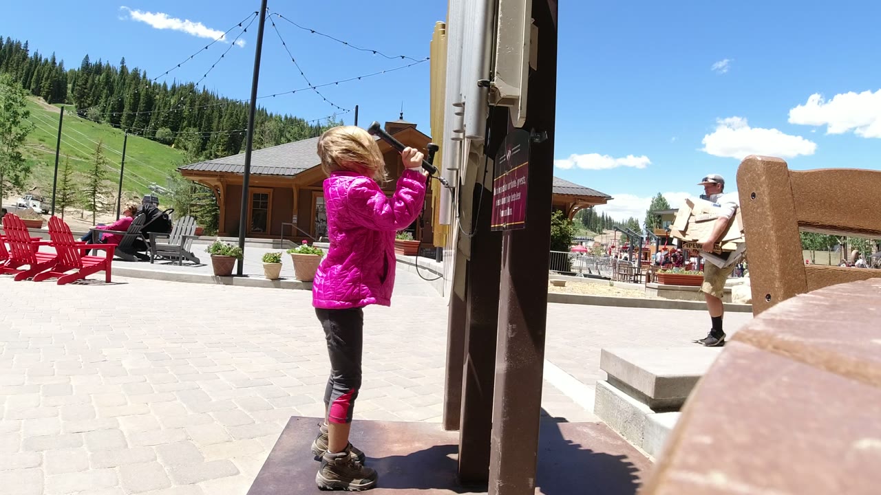 The girl raises one stick toward the sideboard in Winter Park, Colorado, while red chairs, patio tables, a wood building, hillside grass, and trees fill the background.