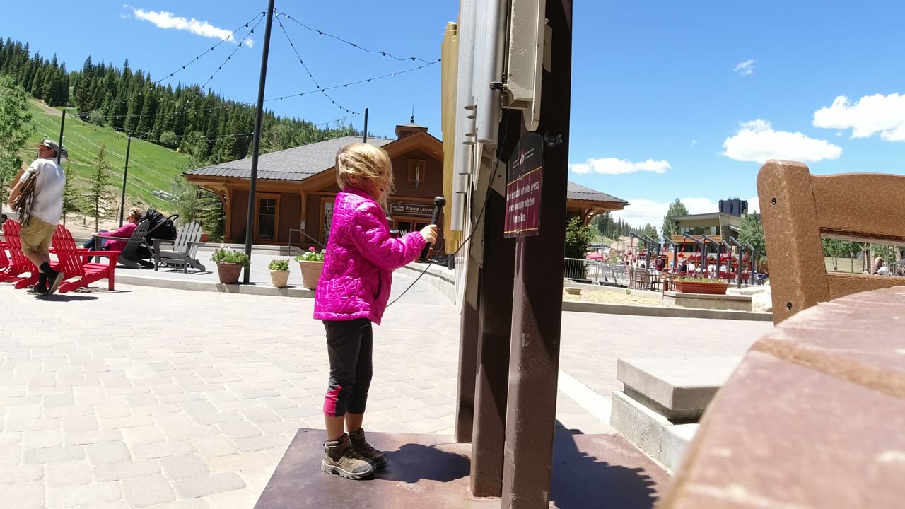 Young girl in a pink jacket standing beside a post in a sunny outdoor plaza.
