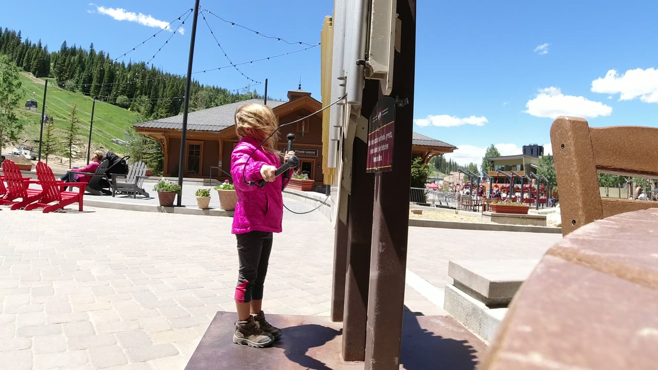 The girl holds the old phone handset out from the post in Winter Park, Colorado, while red chairs, a stroller, wood buildings, string lights, and ski-slope greenery sit behind her.