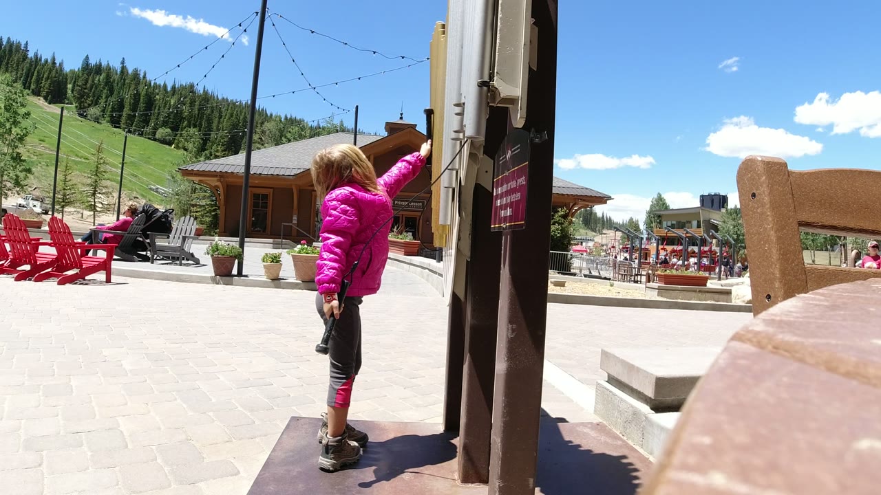 A child in a pink jacket stands beside a dark post in Winter Park, Colorado, with red chairs, a lodge-style building, and a green hillside behind.