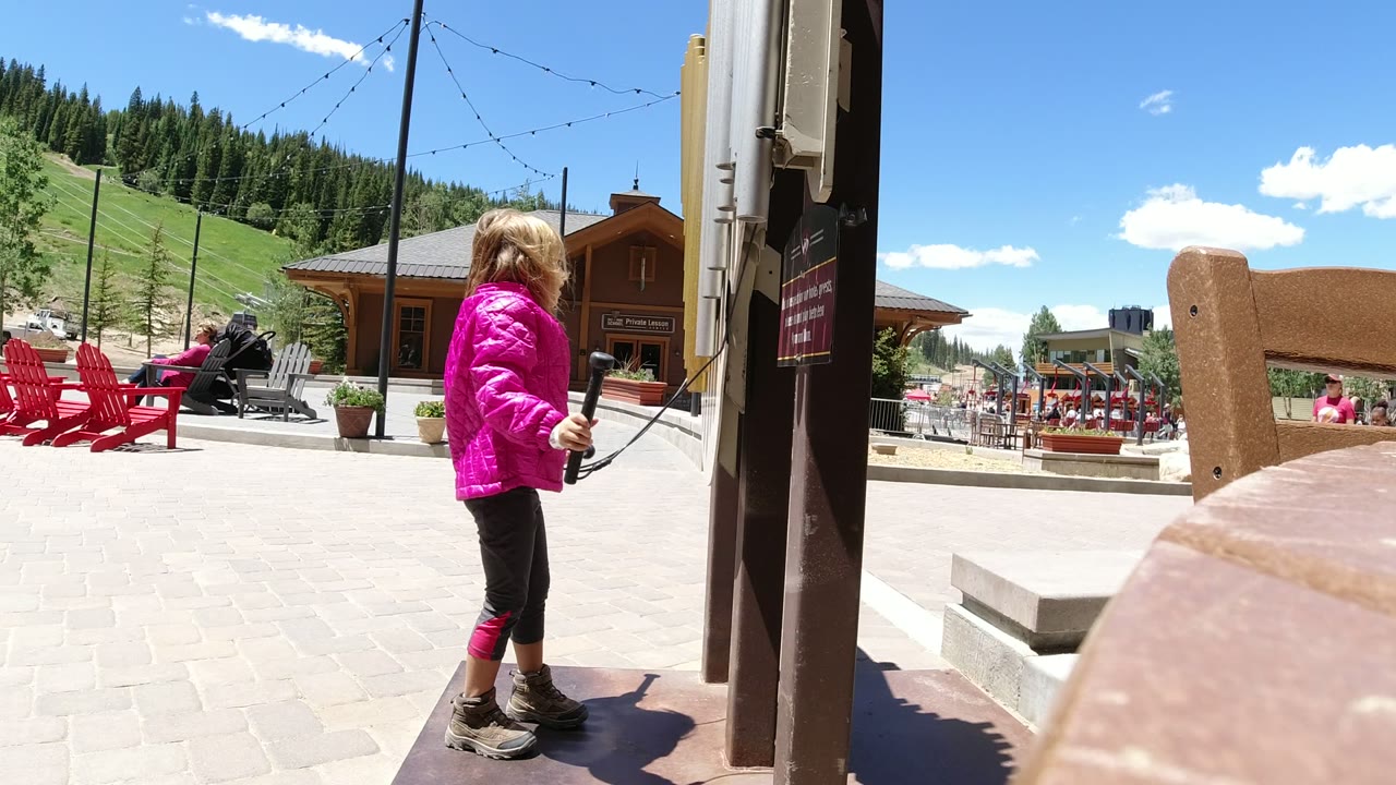 The child holds a hanging striker near the vertical post in Winter Park, Colorado, while red outdoor seating and a wooden building sit across the plaza.