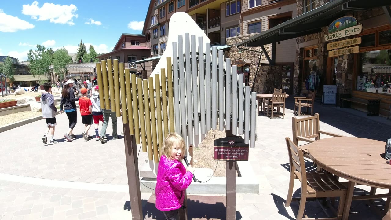 Girl in a pink jacket beside tall metal chimes in a sunny plaza.